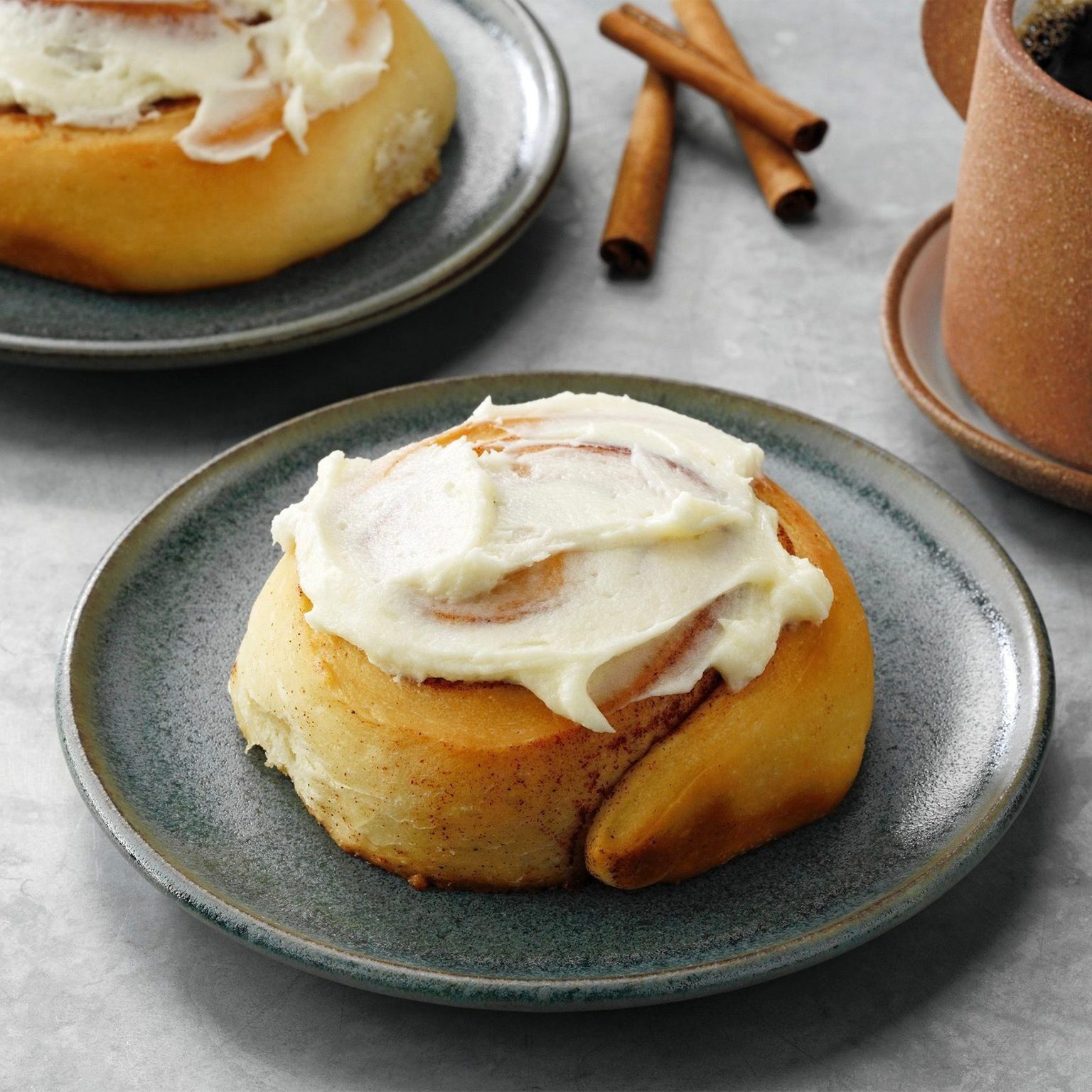 Cinnamon roll rests on a blue-gray plate, topped with white icing; cinnamon sticks and coffee cup in the background.