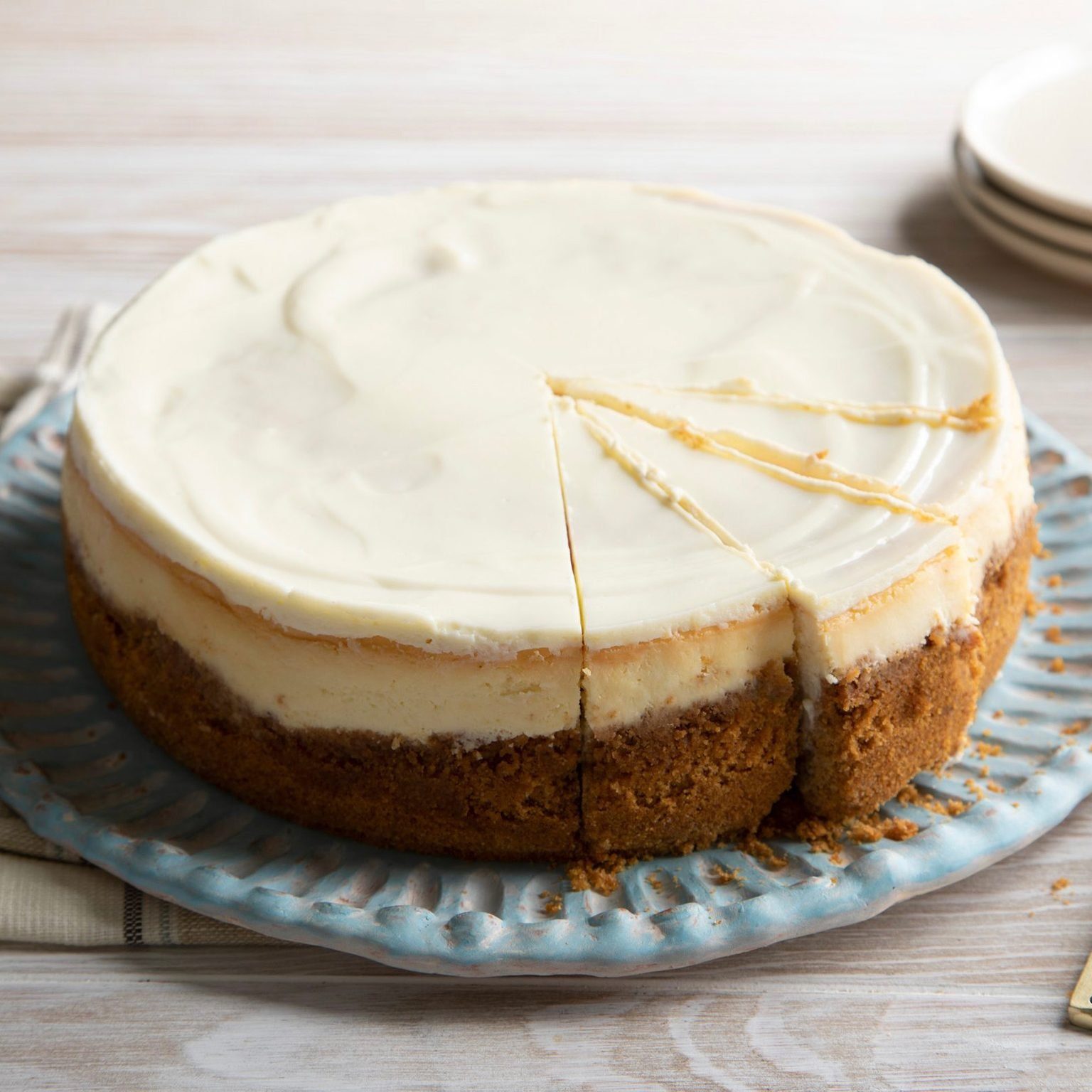 Cheesecake with creamy top and golden crust, partially sliced, rests on a patterned blue plate on a light wooden table.