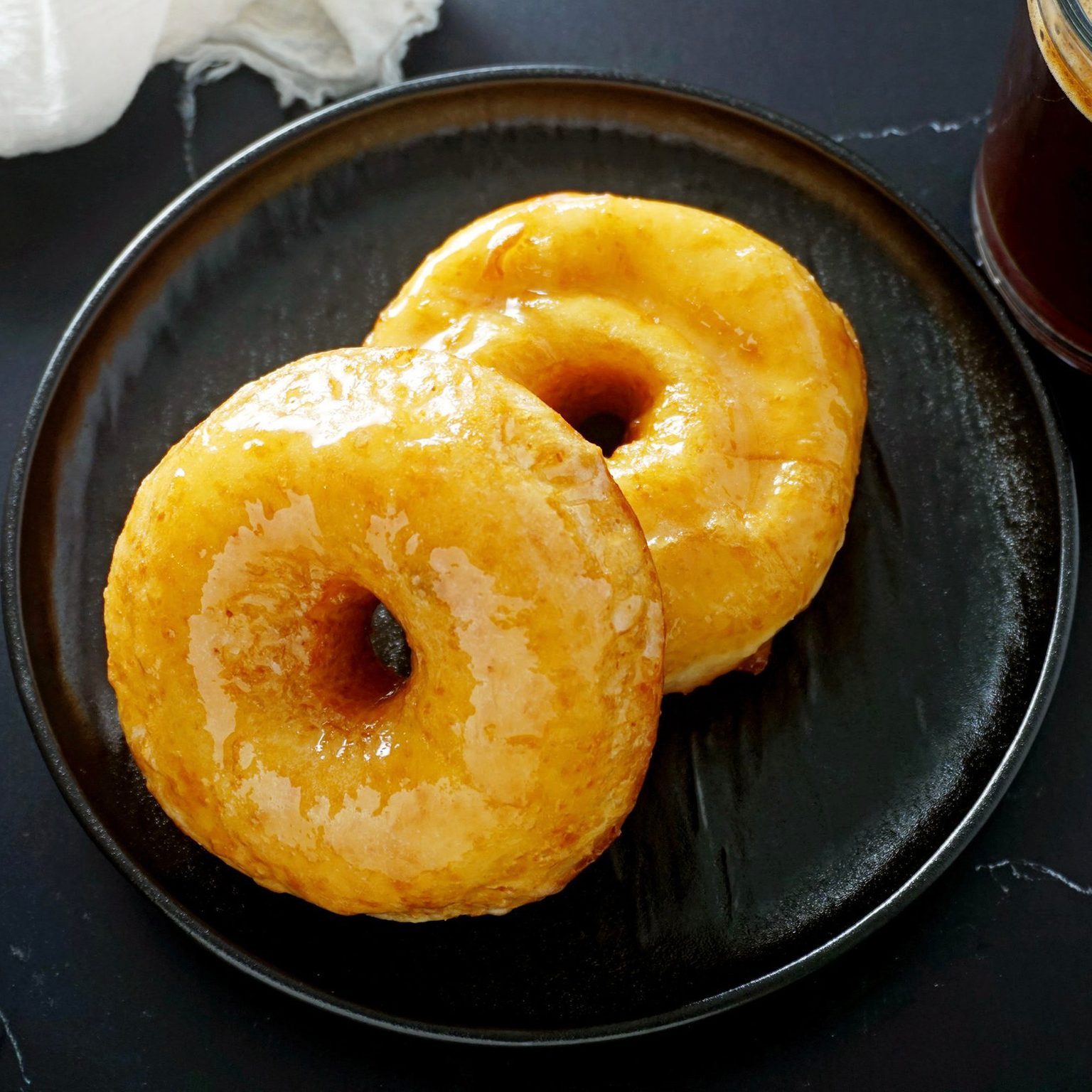 Two glazed donuts rest side-by-side on a black plate, with a dark table surface and a partial coffee cup visible nearby.