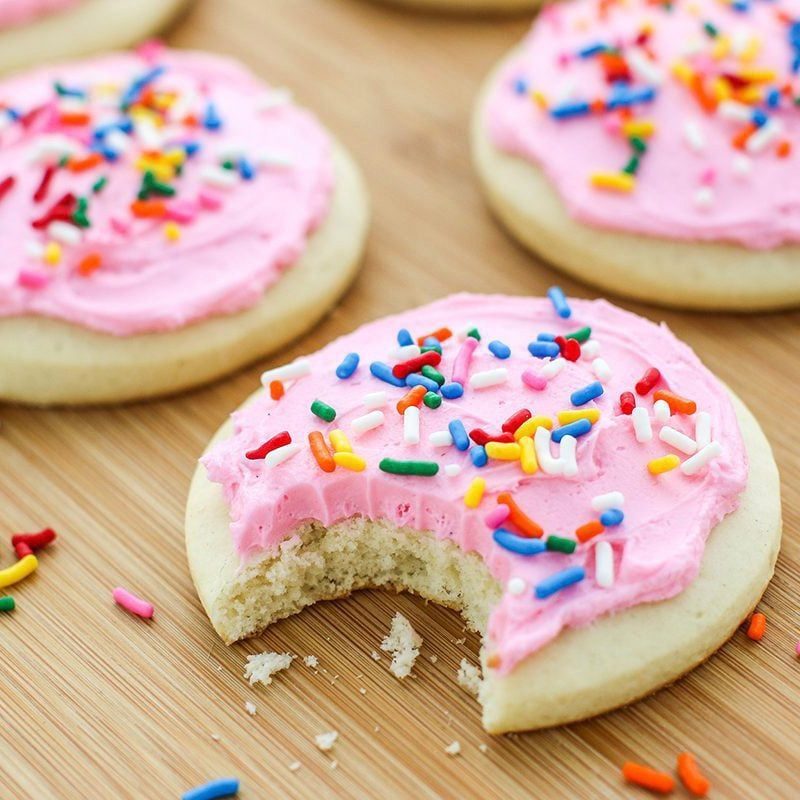 Cookie with pink frosting and sprinkles, partly eaten on a wooden surface. Other similar cookies and colorful sprinkles are scattered around.