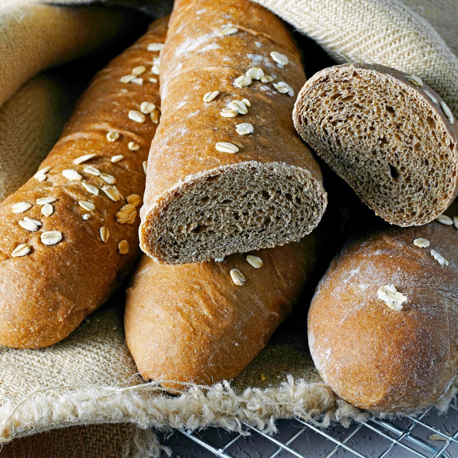Whole grain baguettes rest on burlap, topped with oats, one sliced revealing texture, laid on a cooling rack.