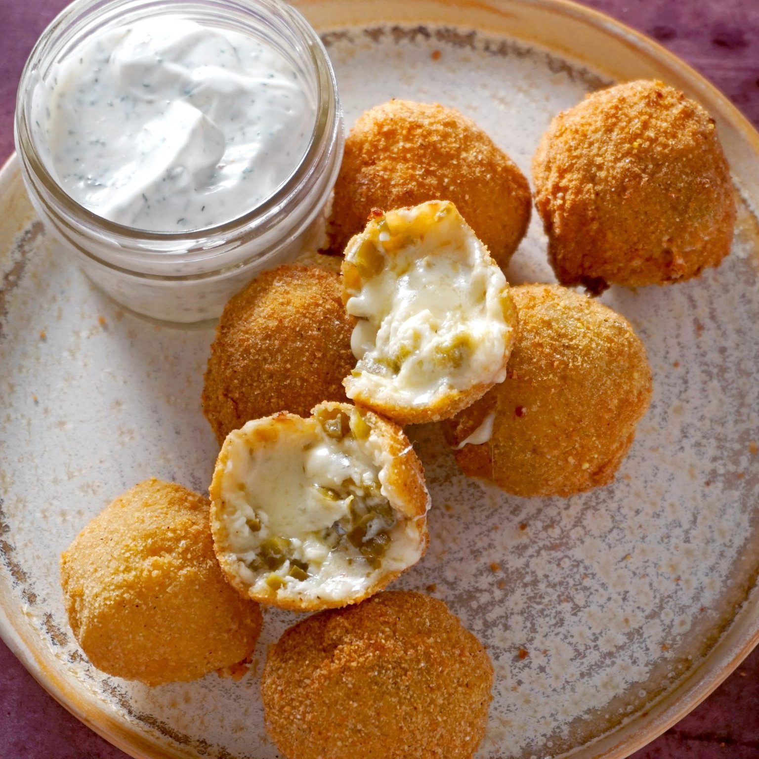 Fried balls with melted cheese filling rest on a plate beside a jar of creamy dipping sauce, on a rustic tabletop.