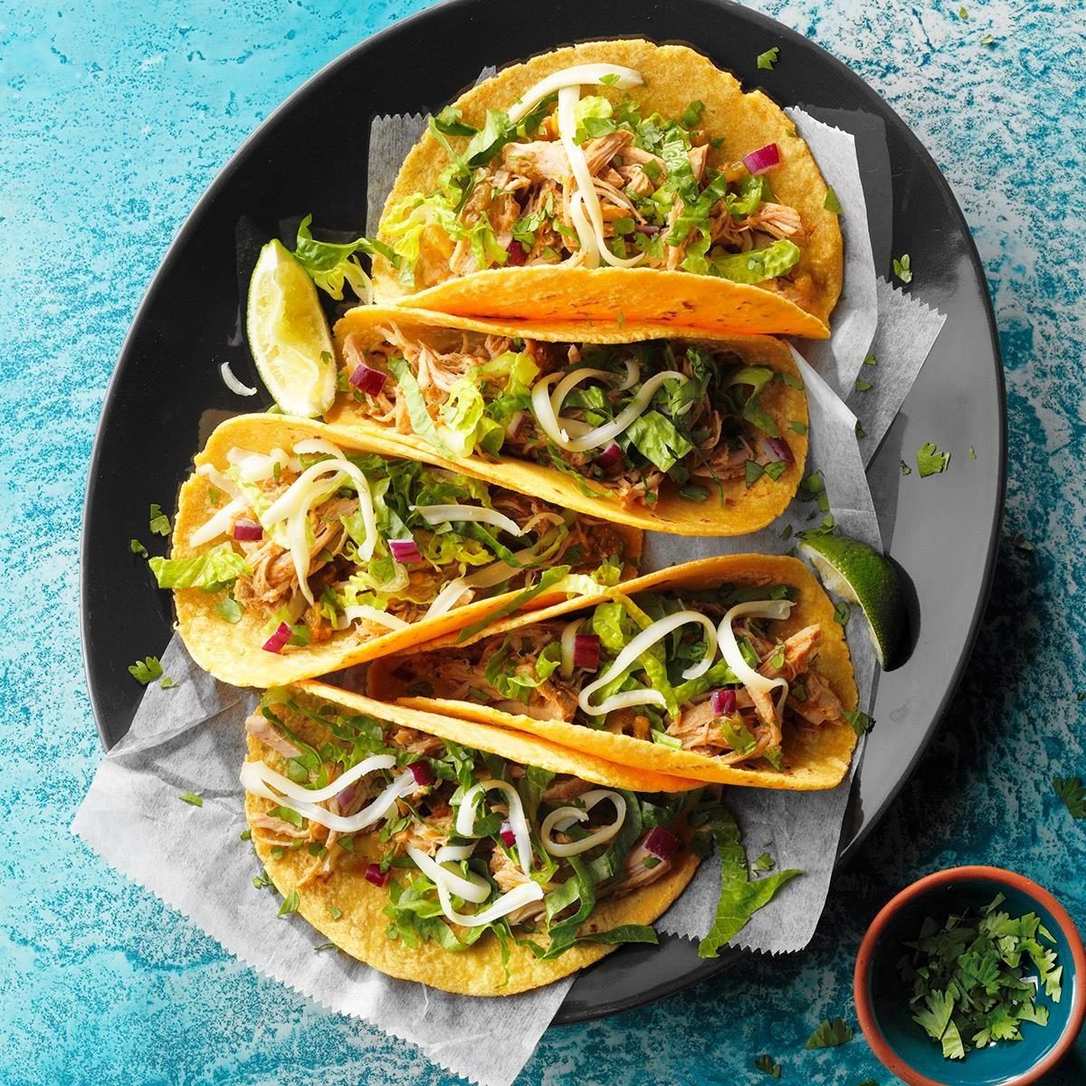 Tacos filled with shredded meat, lettuce, cheese, and onions rest on a dark plate with lime wedges; cilantro is nearby on a blue background.