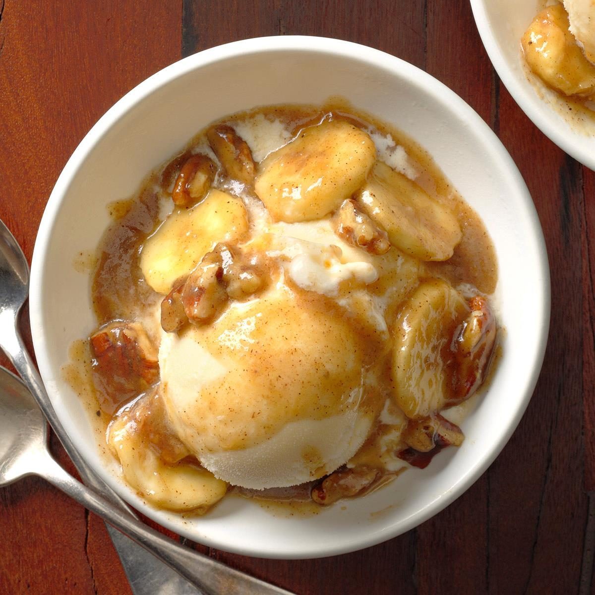 Ice cream topped with caramelized banana slices and nuts, served in a white bowl on a wooden table with two spoons beside it.