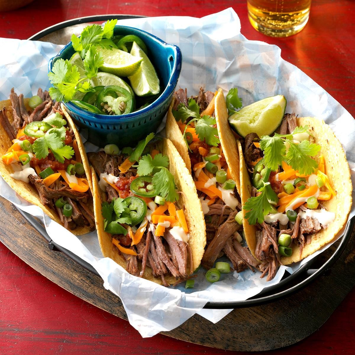 Tacos filled with shredded beef, cheese, and cilantro rest on a tray with lime wedges and jalapeÃ±os in a small blue bowl nearby.