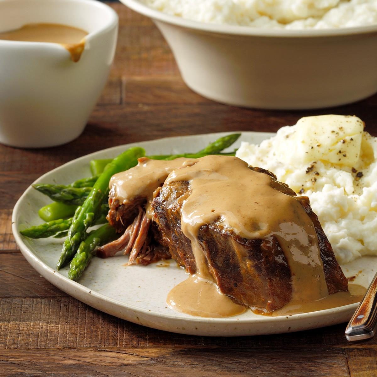Plate holds roast beef with gravy, asparagus, and mashed potatoes; wooden table background.