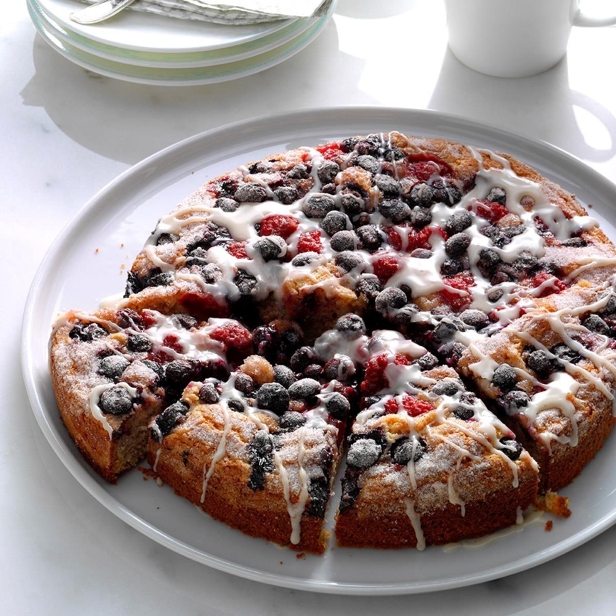Cake topped with mixed berries and icing sits sliced on a white plate, surrounded by a cup and stacked dishes in bright daylight.