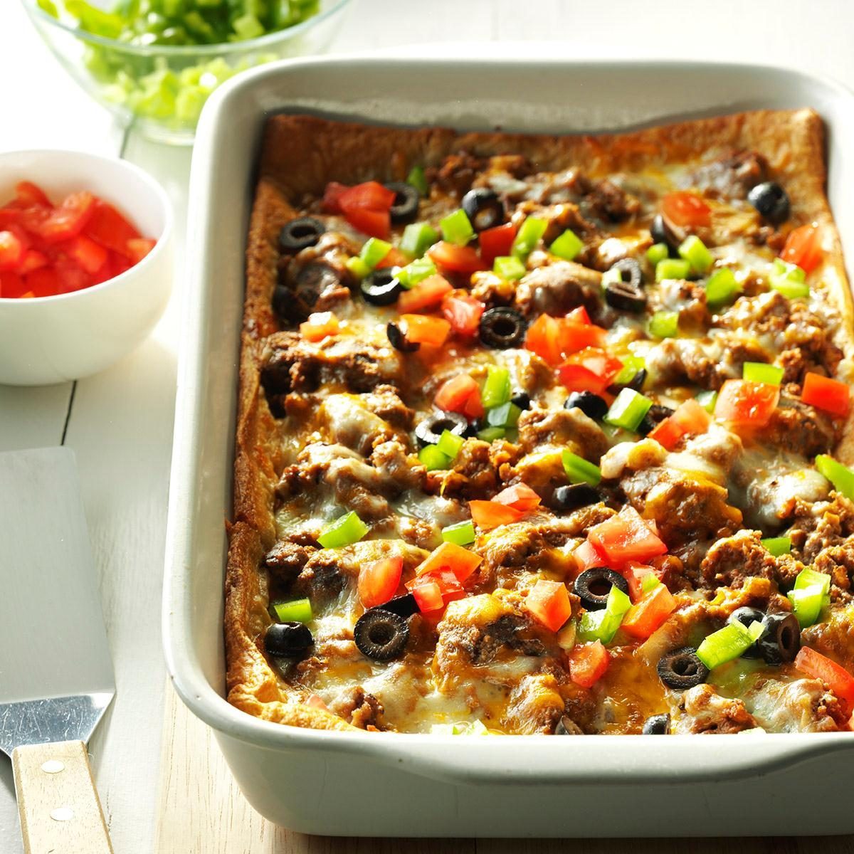 Casserole sits in baking dish, topped with melted cheese, ground meat, diced tomatoes, green peppers, and olives; extra toppings in bowls nearby.