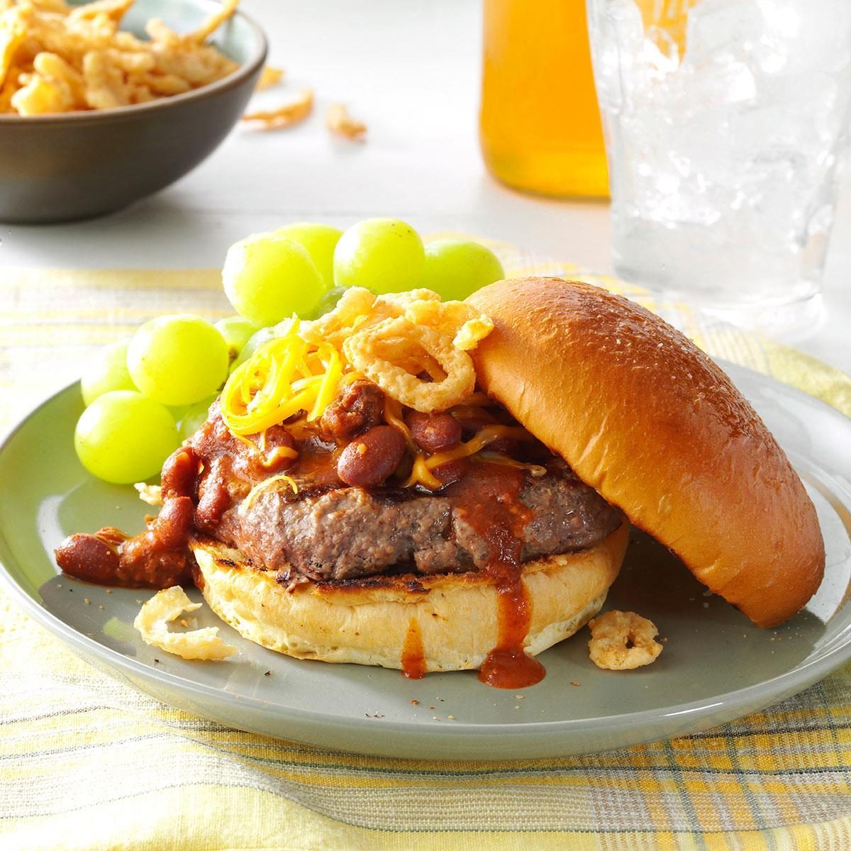 Burger topped with chili, cheese, and onions sits on a green plate with green grapes; tablecloth and drink in the background.