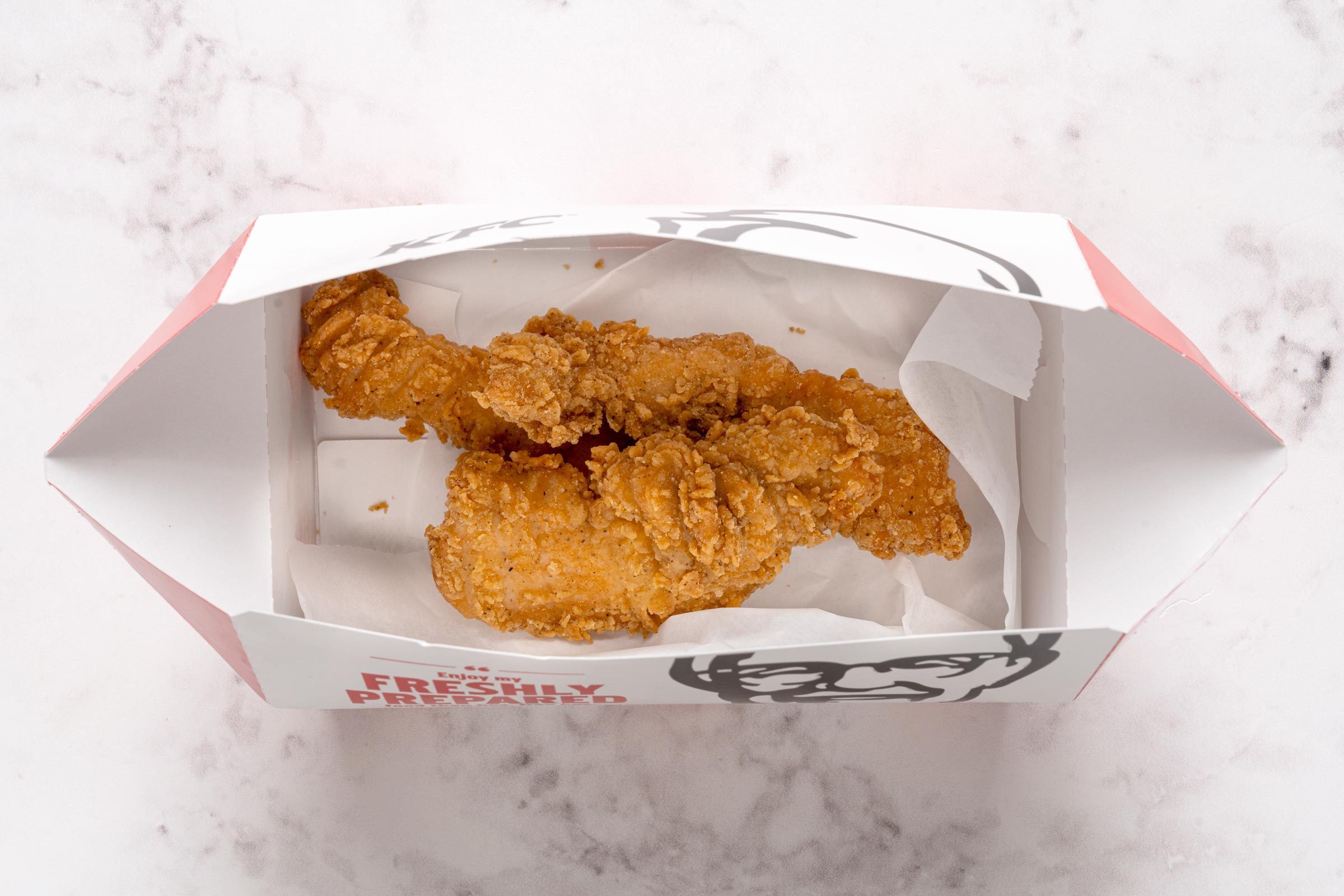 Fried chicken pieces rest in an open KFC box on a light marbled surface.