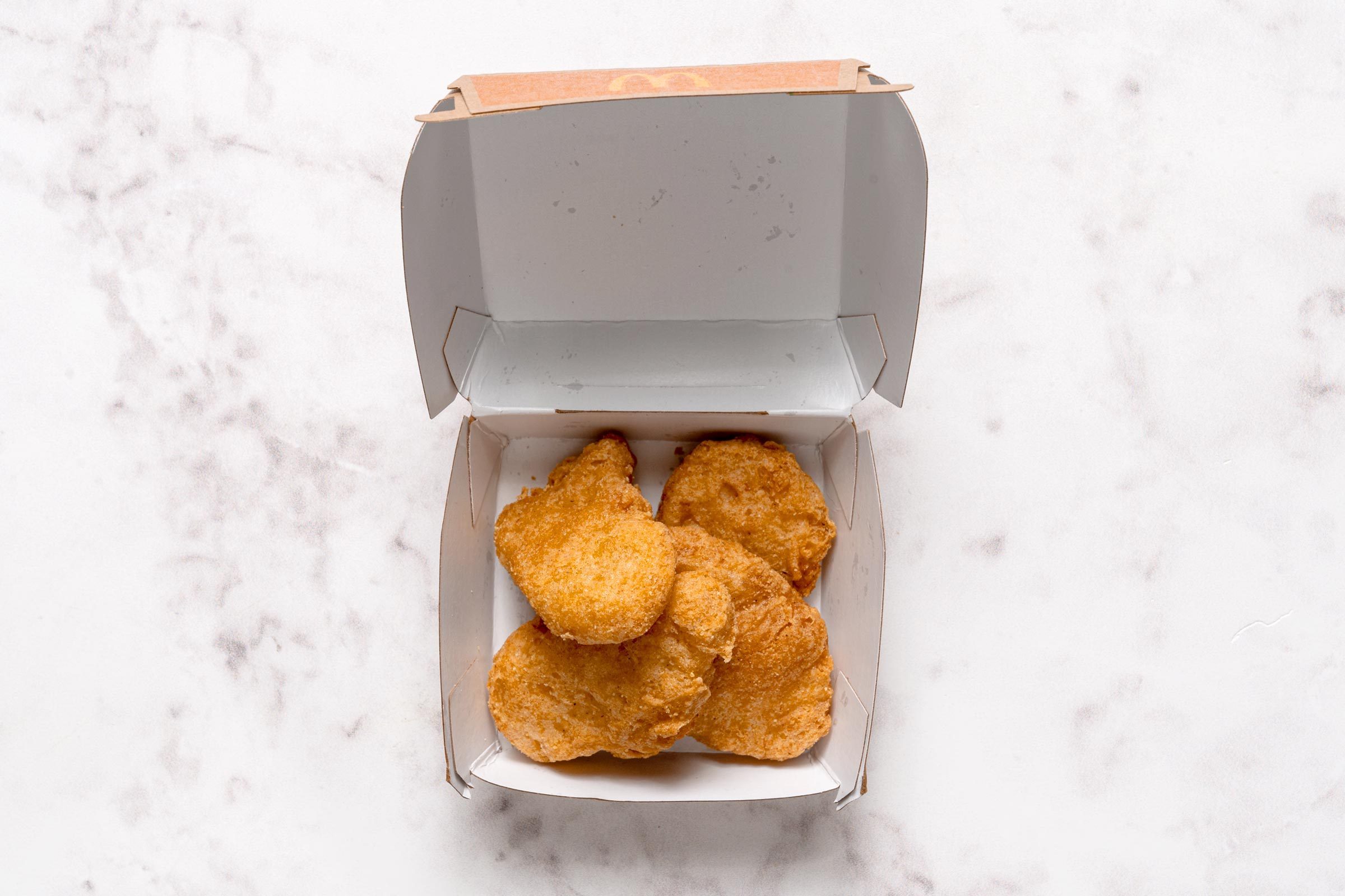 Four chicken nuggets sit inside an open cardboard box on a white marbled surface.