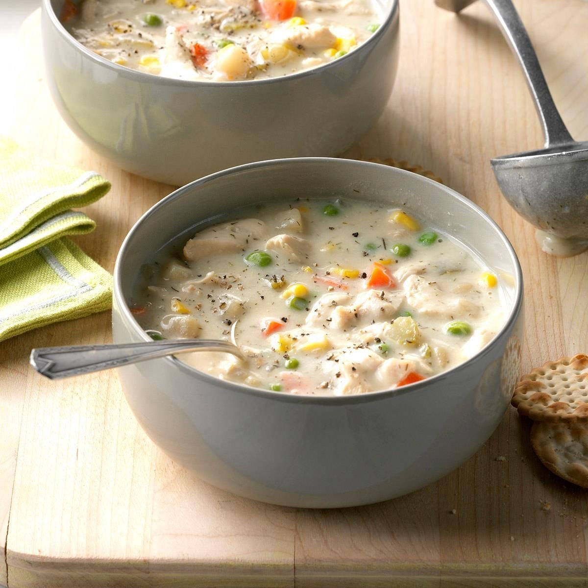 Creamy soup sits in a gray bowl with vegetables and chicken, surrounded by a ladle, crackers, and a green cloth on a wooden surface.