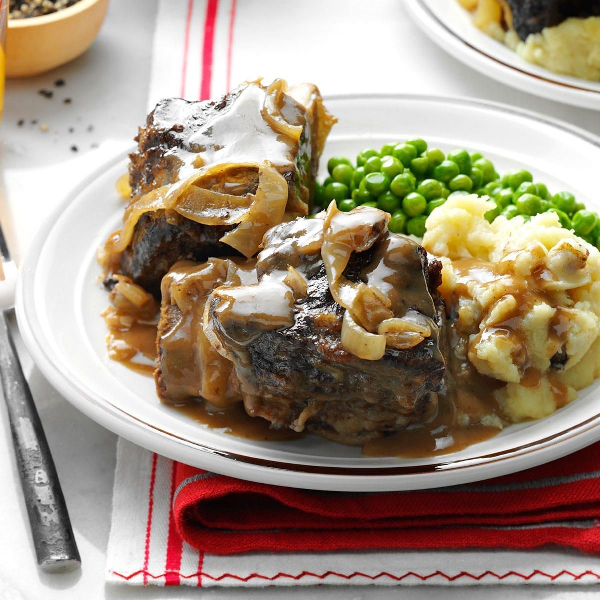 Beef with onions is covered in gravy, sitting on a plate alongside mashed potatoes and peas, placed on a red-striped napkin.