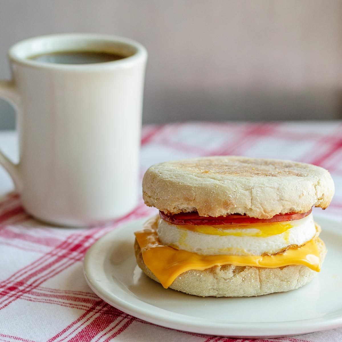 Breakfast sandwich sits on a plate, next to a mug filled with coffee, on a red-and-white checkered tablecloth.