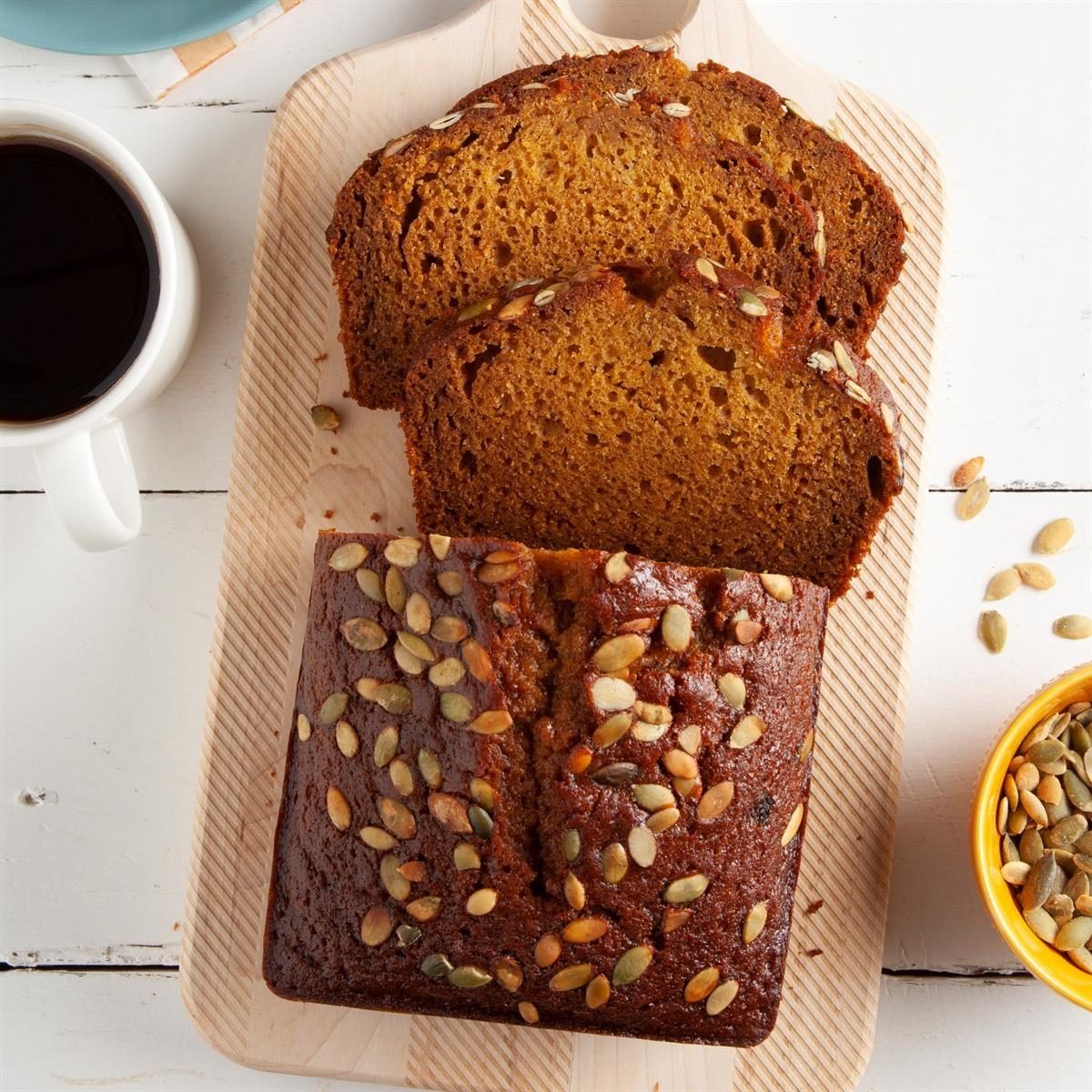Sliced pumpkin bread with seeds rests on a wooden board; a cup of coffee and a bowl of seeds sit nearby on a white table.