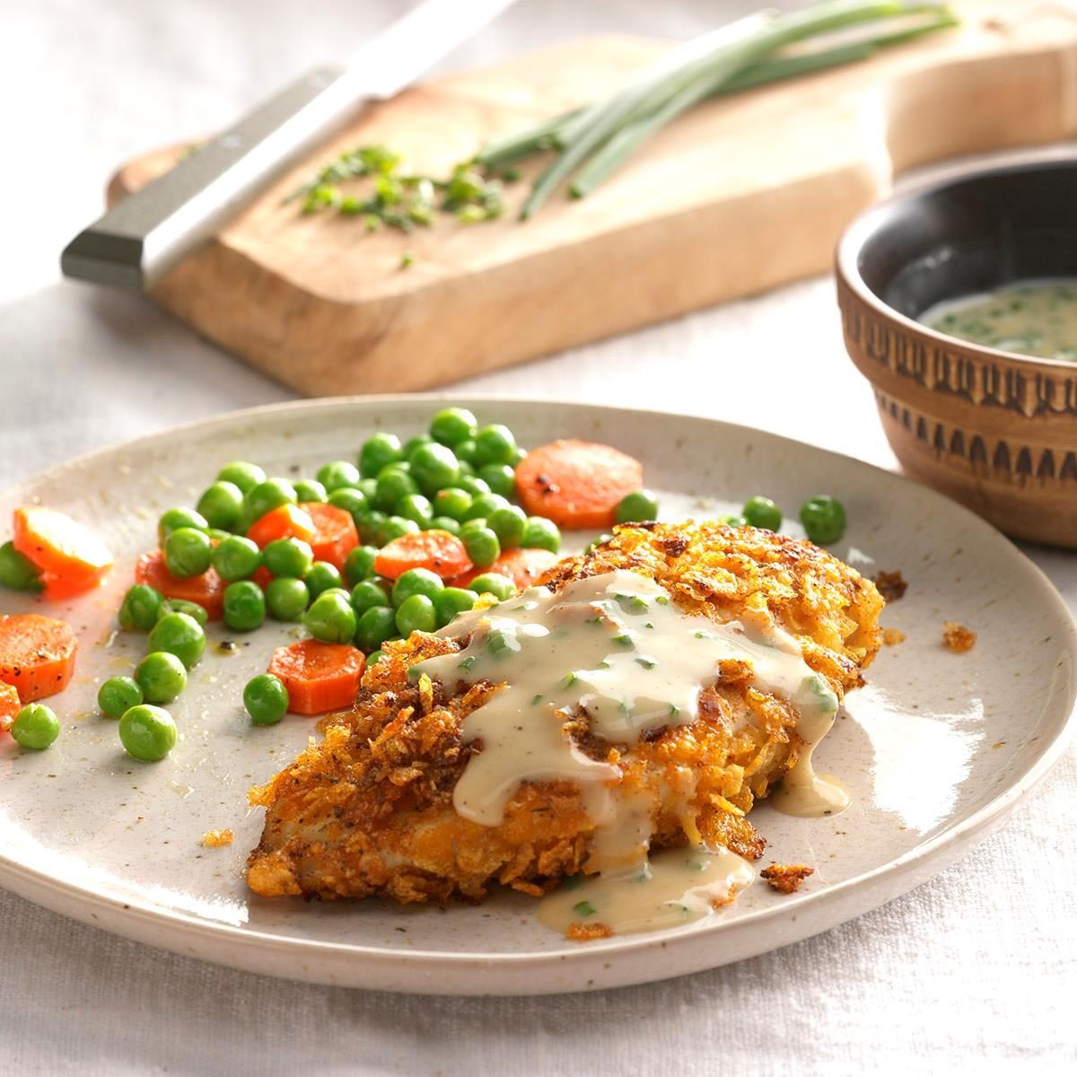 Breaded chicken with creamy sauce rests on a plate with peas and carrots; a wooden board with herbs and a bowl are nearby.