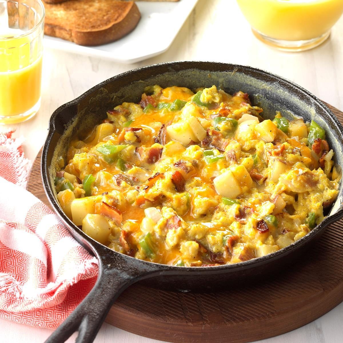 Skillet holds scrambled eggs with vegetables and cheese, resting on a wooden board. Nearby, orange juice and toasted bread enhance the breakfast setting.