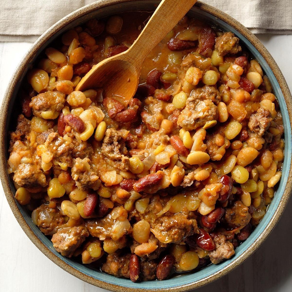 A bowl contains a mix of cooked beans and meat, stirred with a wooden spoon, placed on a light wooden table.