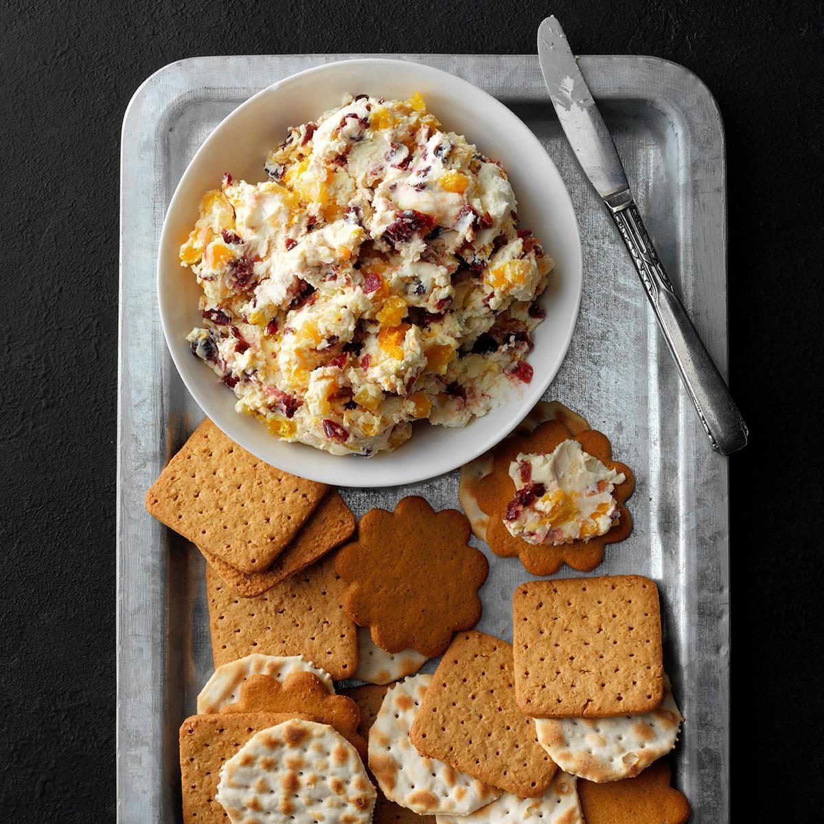 A bowl of fruit-studded cream cheese sits on a metal tray with assorted crackers beside a butter knife.