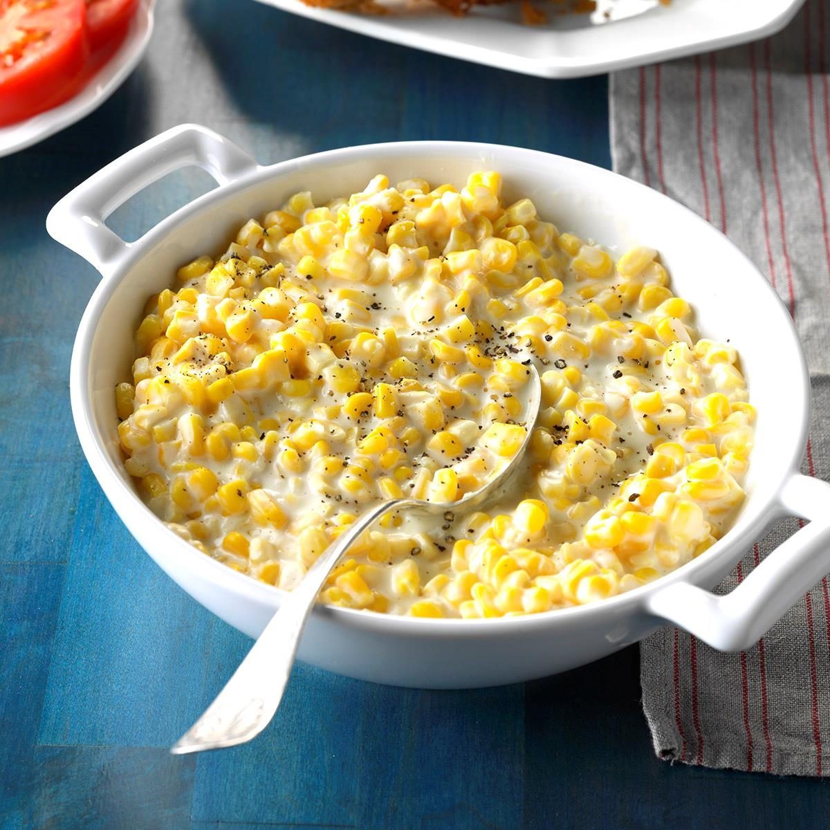 Creamed corn sits in a white dish with a spoon, resting on a blue table beside a striped cloth and sliced tomatoes.