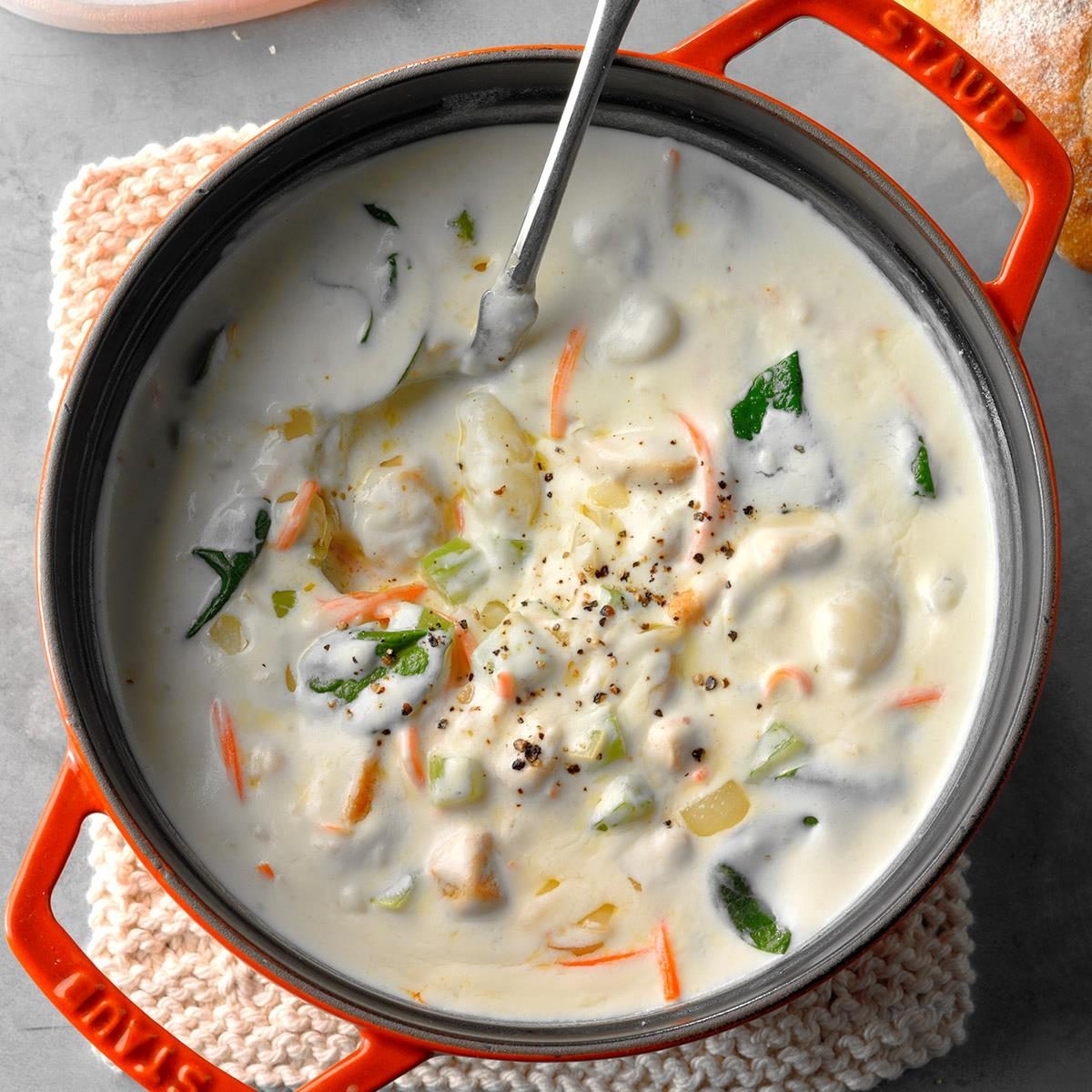 A pot filled with creamy soup and vegetables, resting on a knitted mat, with a spoon inside, on a gray tabletop background.