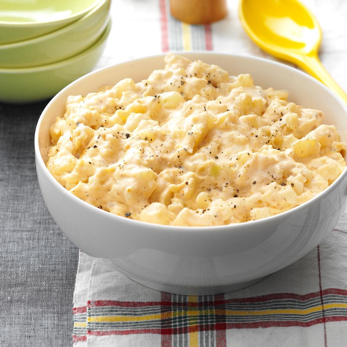 A white bowl holds creamy potato salad, seasoned with pepper, placed on a plaid-patterned cloth beside stacked green bowls and a yellow spoon.