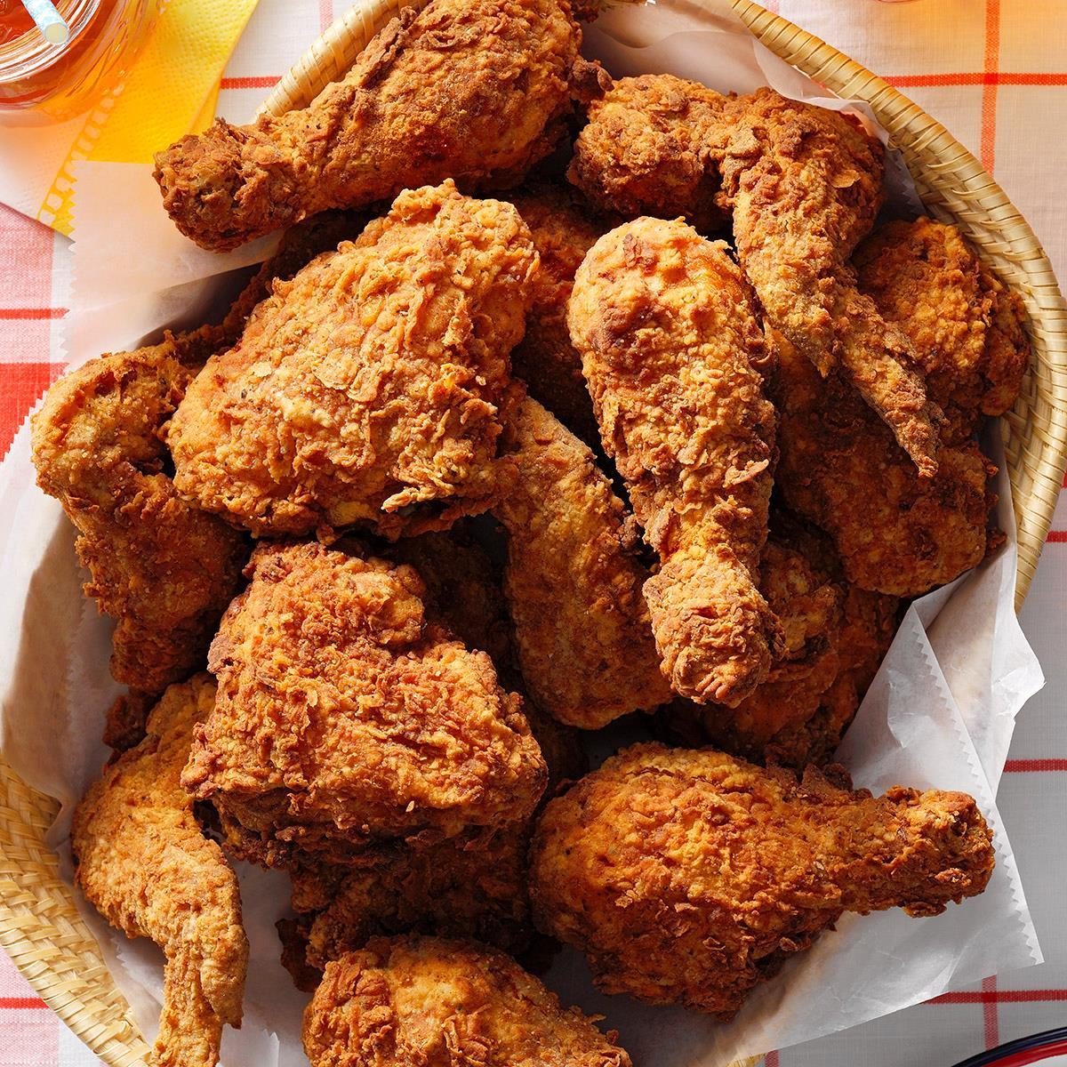 Fried chicken pieces rest in a woven basket lined with paper, placed on a checkered tablecloth with a hint of a honey jar nearby.