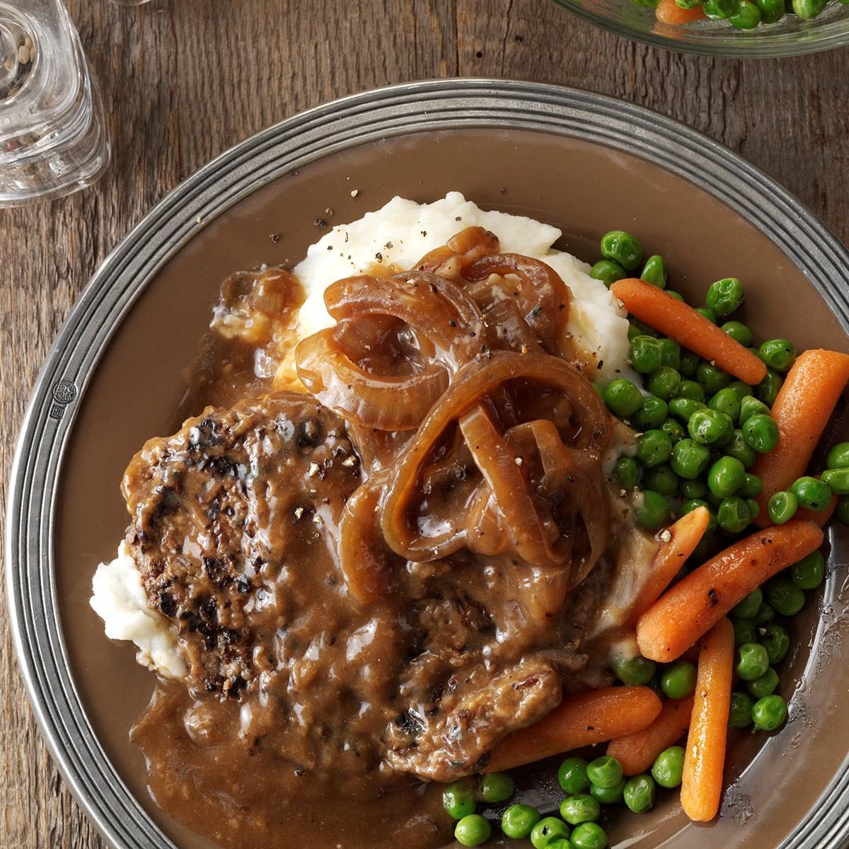 Steak with onion gravy sits on mashed potatoes, accompanied by carrots and peas on a brown plate on a wooden table.