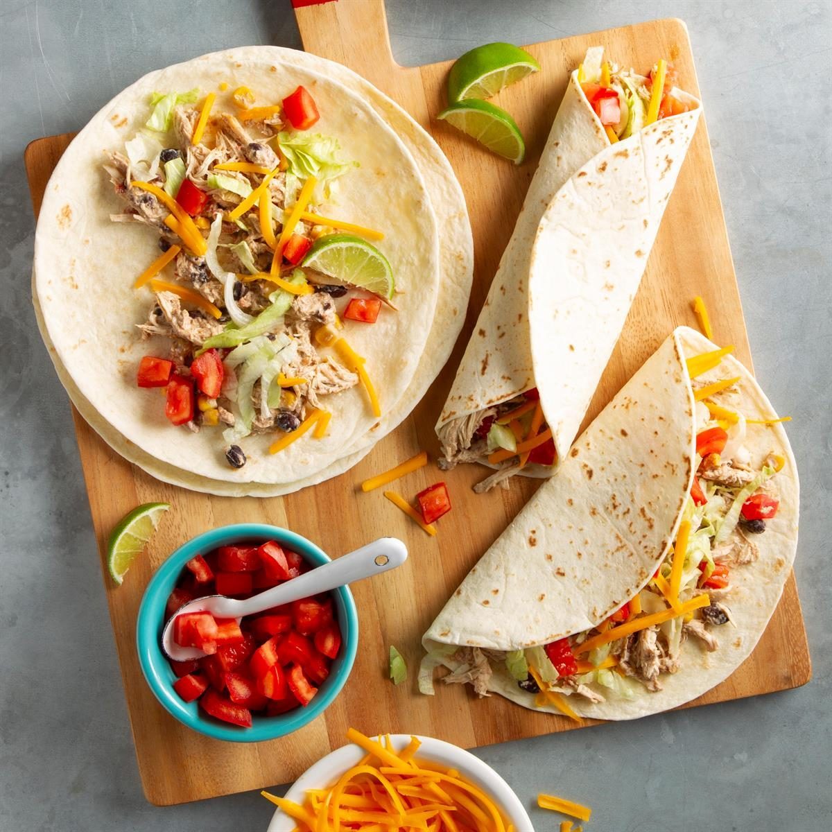 Tortillas filled with chicken, cheese, and vegetables on a wooden board; surrounded by lime wedges, diced tomatoes in a bowl, and shredded cheese.