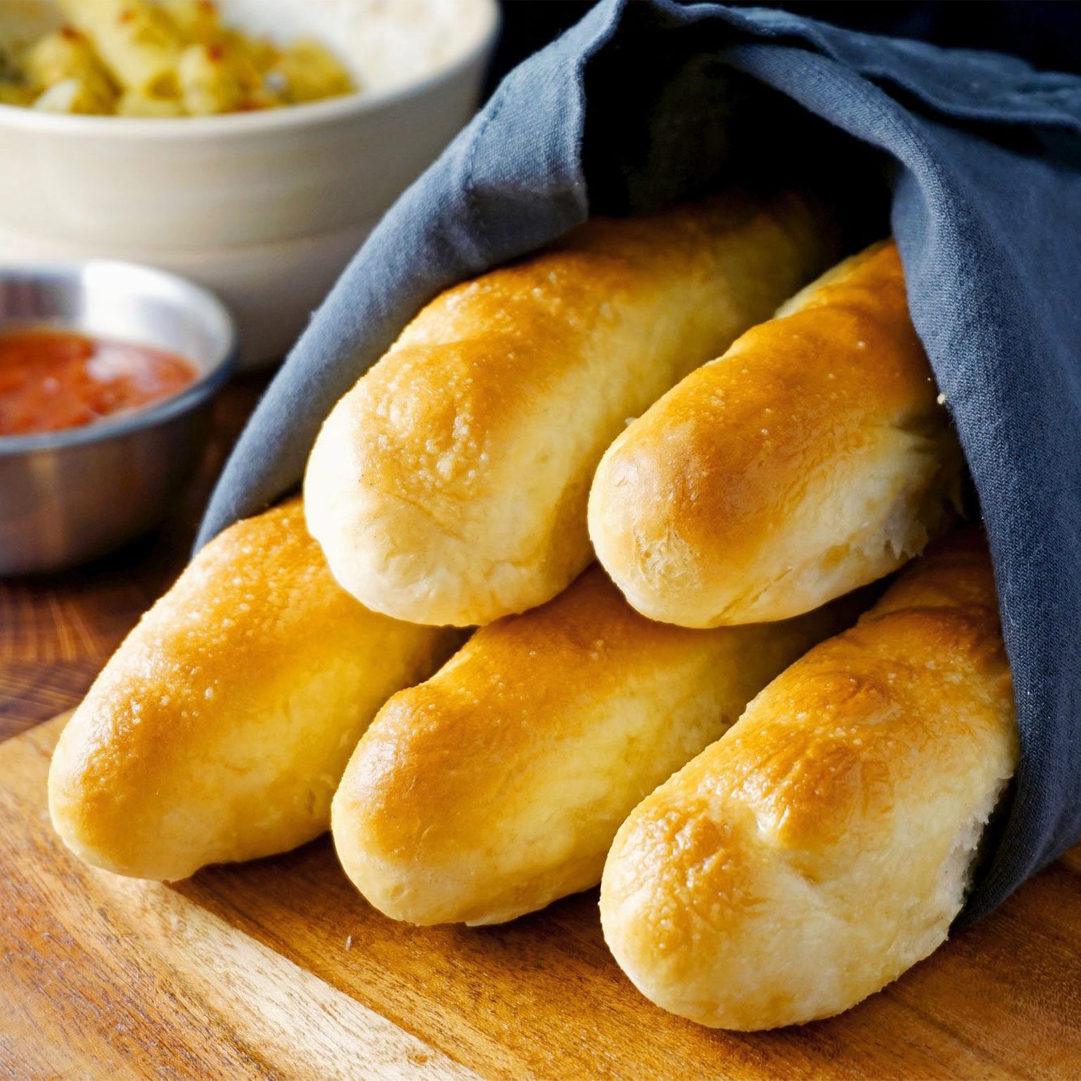Breadsticks wrapped in a dark cloth lie on a wooden board, with dipping sauces in bowls nearby.