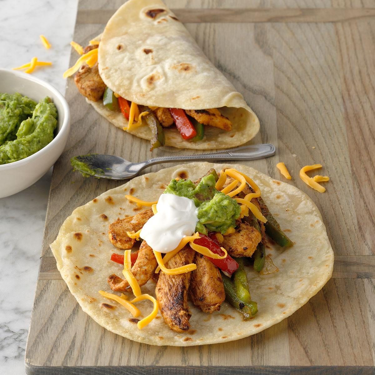 Tortilla holds seasoned chicken, peppers, cheese, guacamole, and sour cream; nearby, another folded tortilla rests; bowl of guacamole and fork accompany on wooden surface.