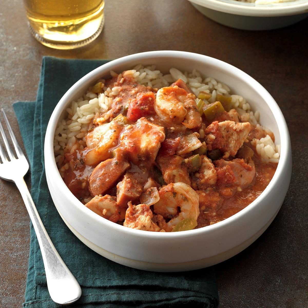 A white bowl holds gumbo served over rice, placed on a dark cloth with a fork nearby, beside a glass of amber liquid.
