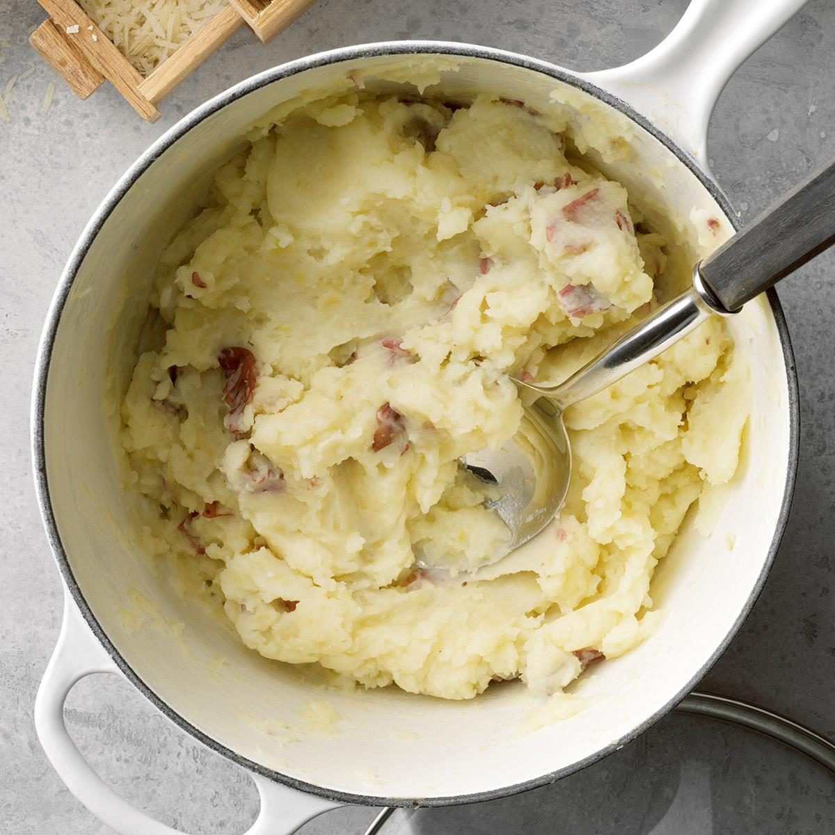 Mashed potatoes rest in a white pot with a metal spoon, placed on a gray surface beside a wooden cheese grater.