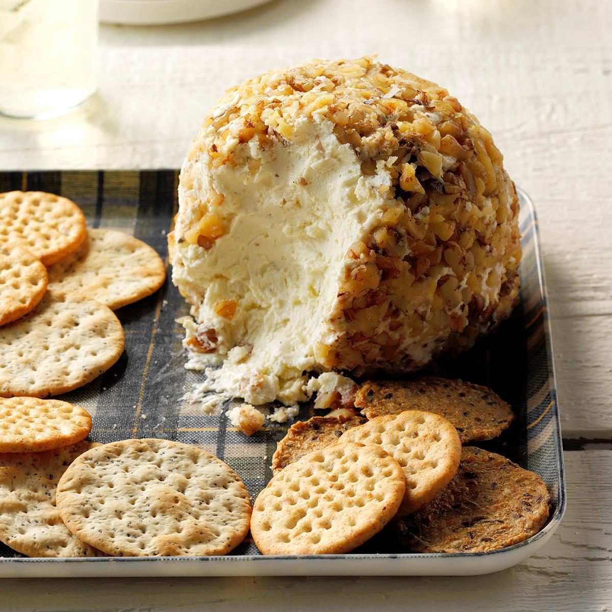 Cheese ball with nuts partially eaten, surrounded by assorted crackers, placed on a checkered tray atop a wooden table.