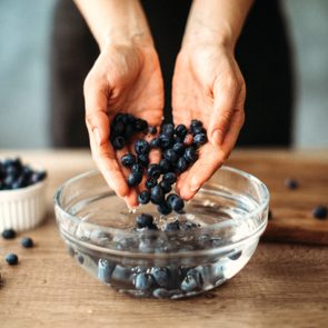 Hands gently drop blueberries into clear bowl of water on wooden table.