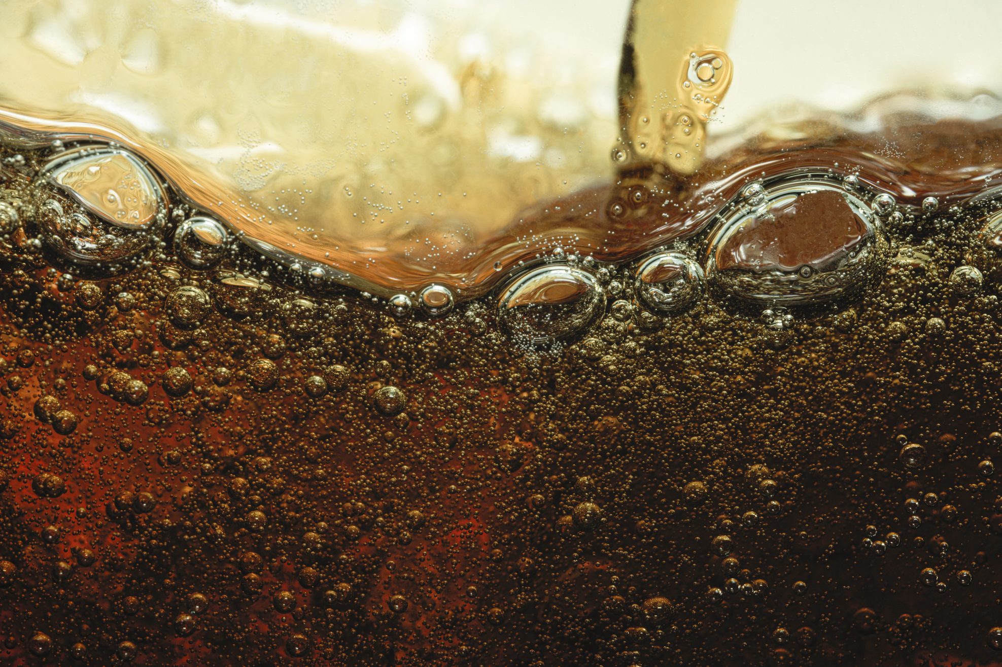 Bubbles rise in a dark, fizzy liquid being poured, creating a foamy surface in close-up view.