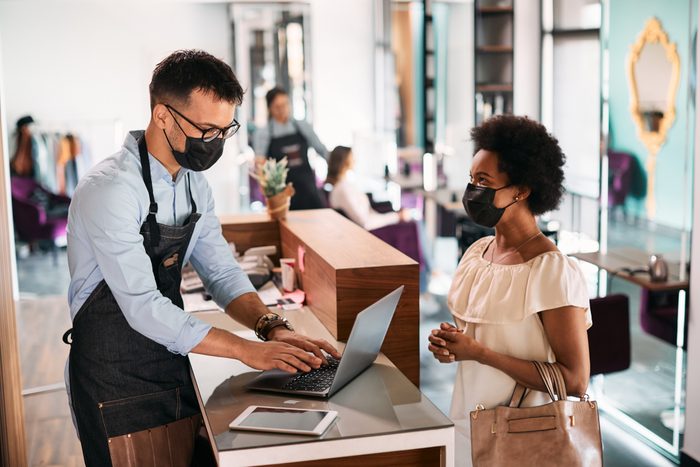 Hairstylist using laptop while talking to African American woman at reception desk.