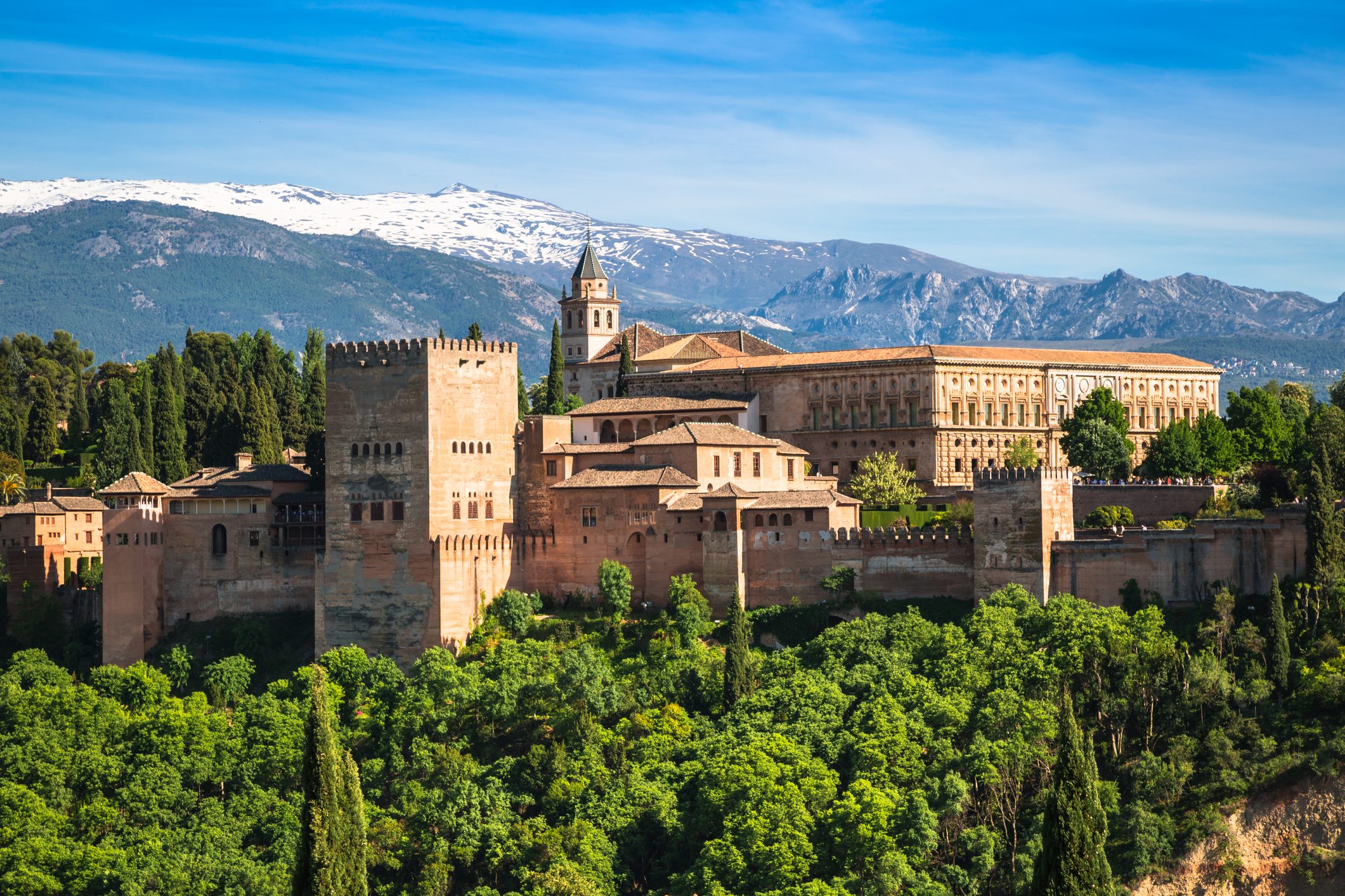 A historic castle stands atop a lush green landscape, with snow-capped mountains in the background under a clear blue sky.