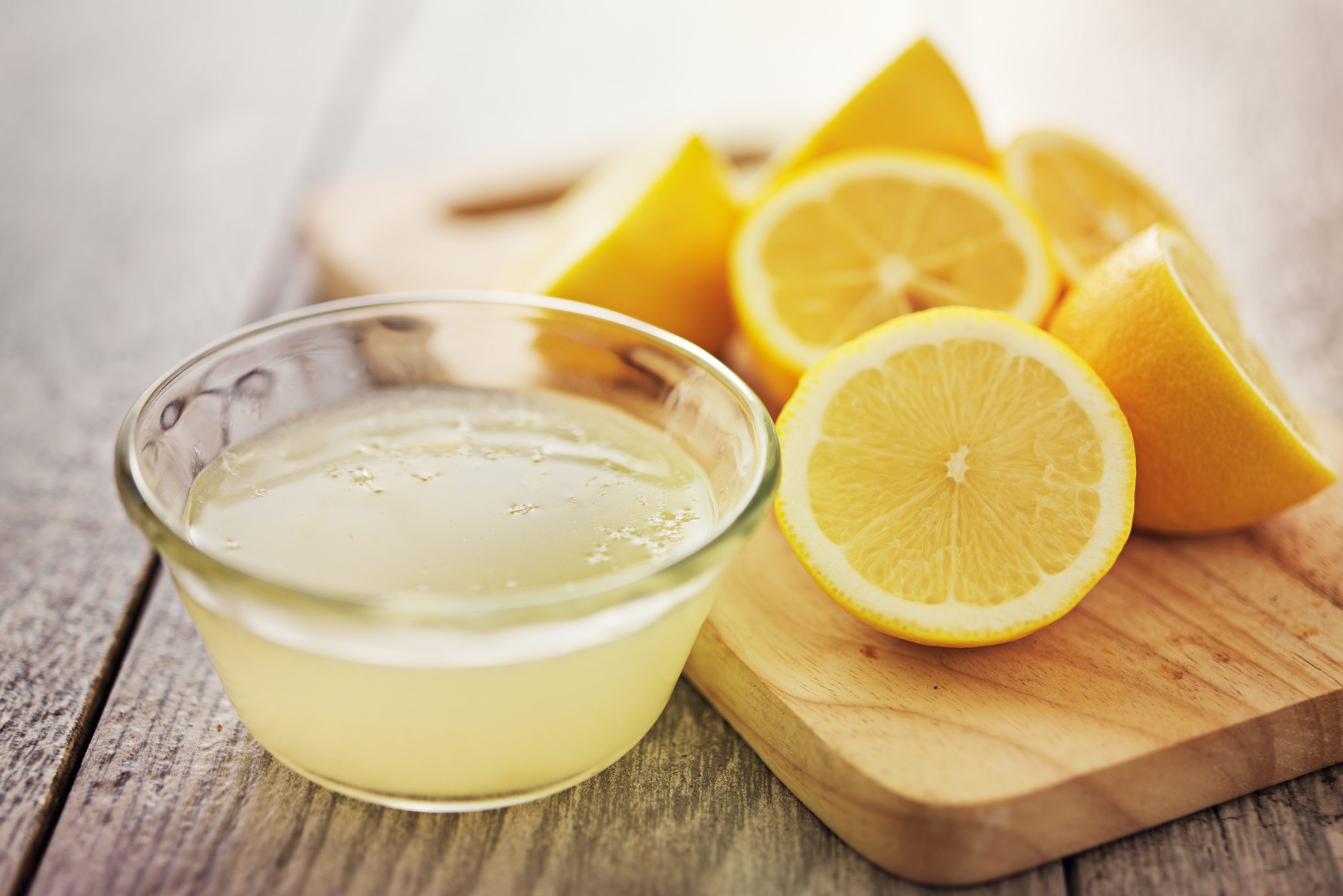 Lemon juice in a glass bowl sits beside sliced lemons on a wooden cutting board on a rustic table.