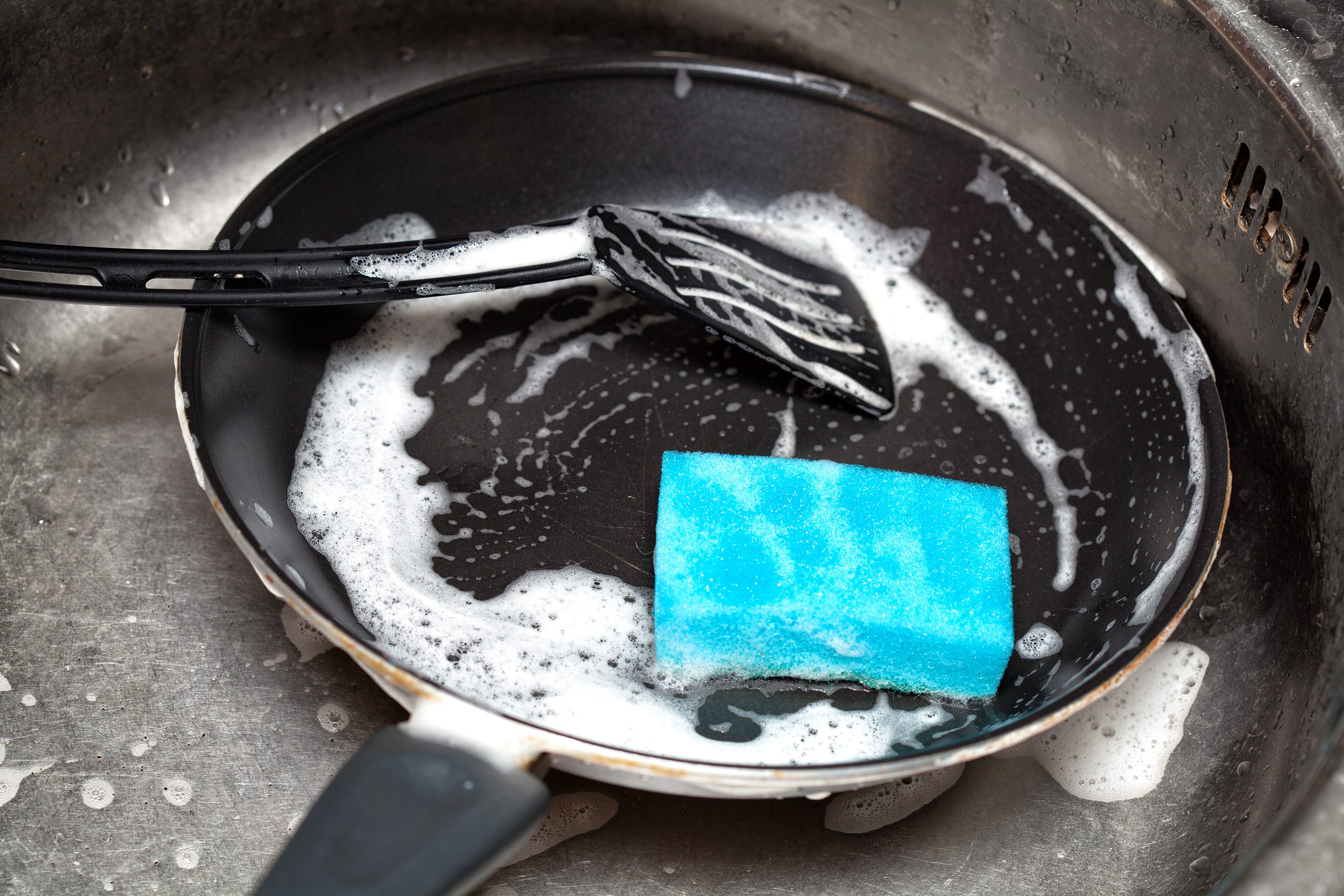 A frying pan with soapy water holds a blue sponge and a spatula, resting in a kitchen sink for cleaning.