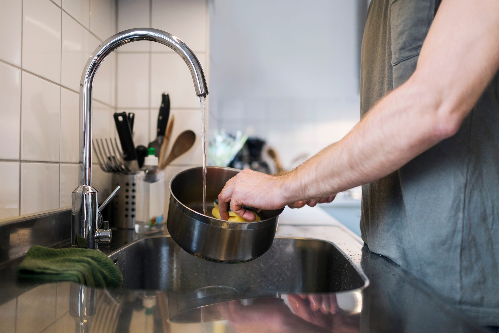 A person holds a pot under running water in a kitchen sink with utensils in the background.