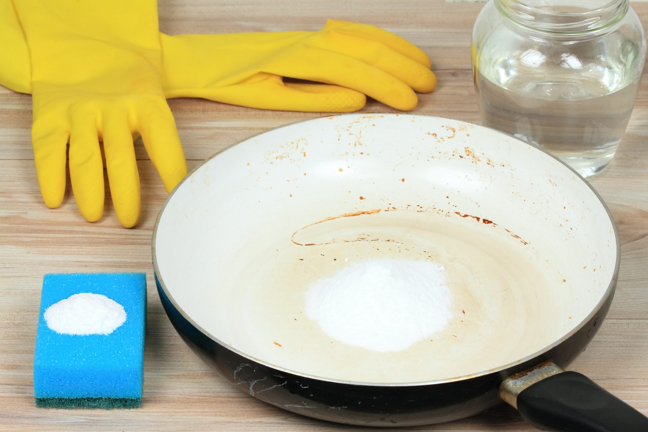 Dirty pan with baking soda, sponge, and water for cleaning; yellow rubber gloves nearby on a wooden surface.