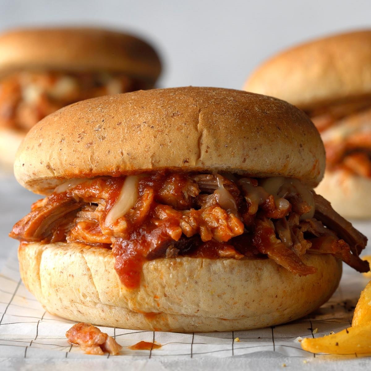 A pulled pork sandwich sits with sauce-drenched meat between buns, on a table with another sandwich nearby, against a neutral background.