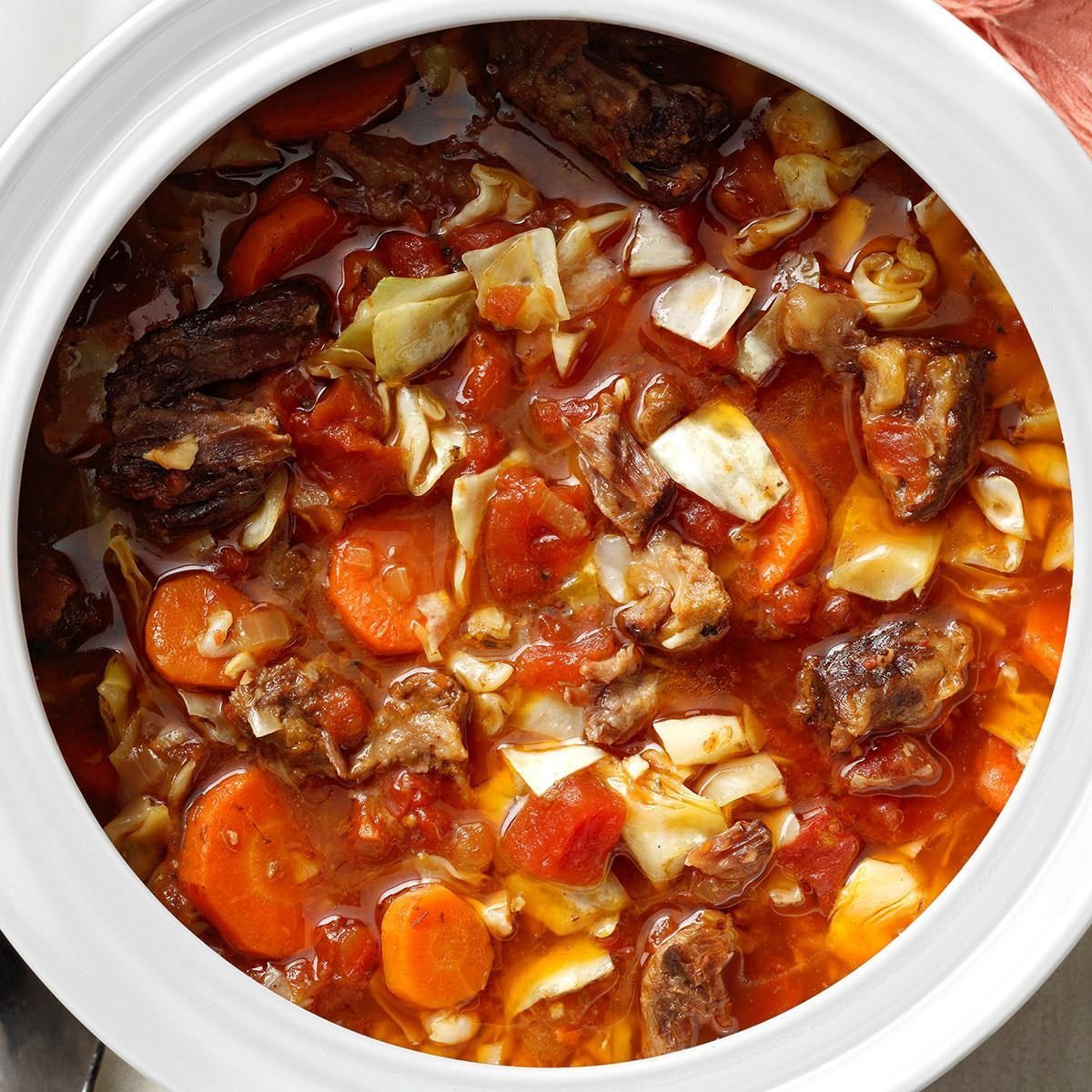 Soup, containing chunks of meat, carrots, and cabbage, is served in a white bowl, resting on a light-colored surface.