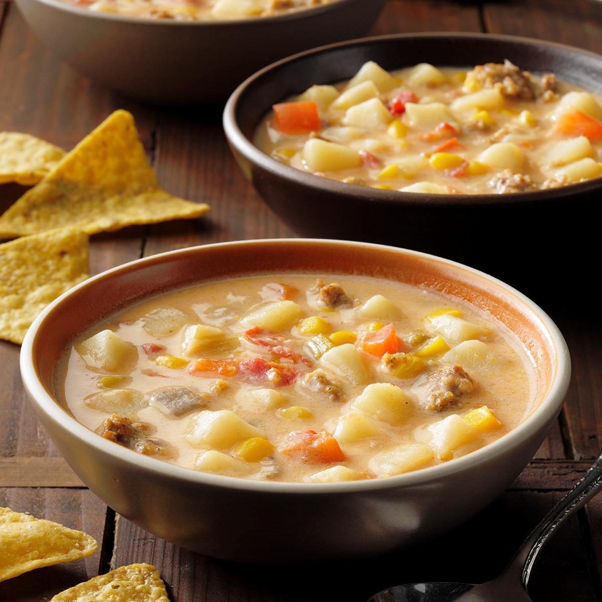 Bowls of creamy soup sit on a wooden table, filled with diced vegetables and meat, accompanied by scattered tortilla chips.