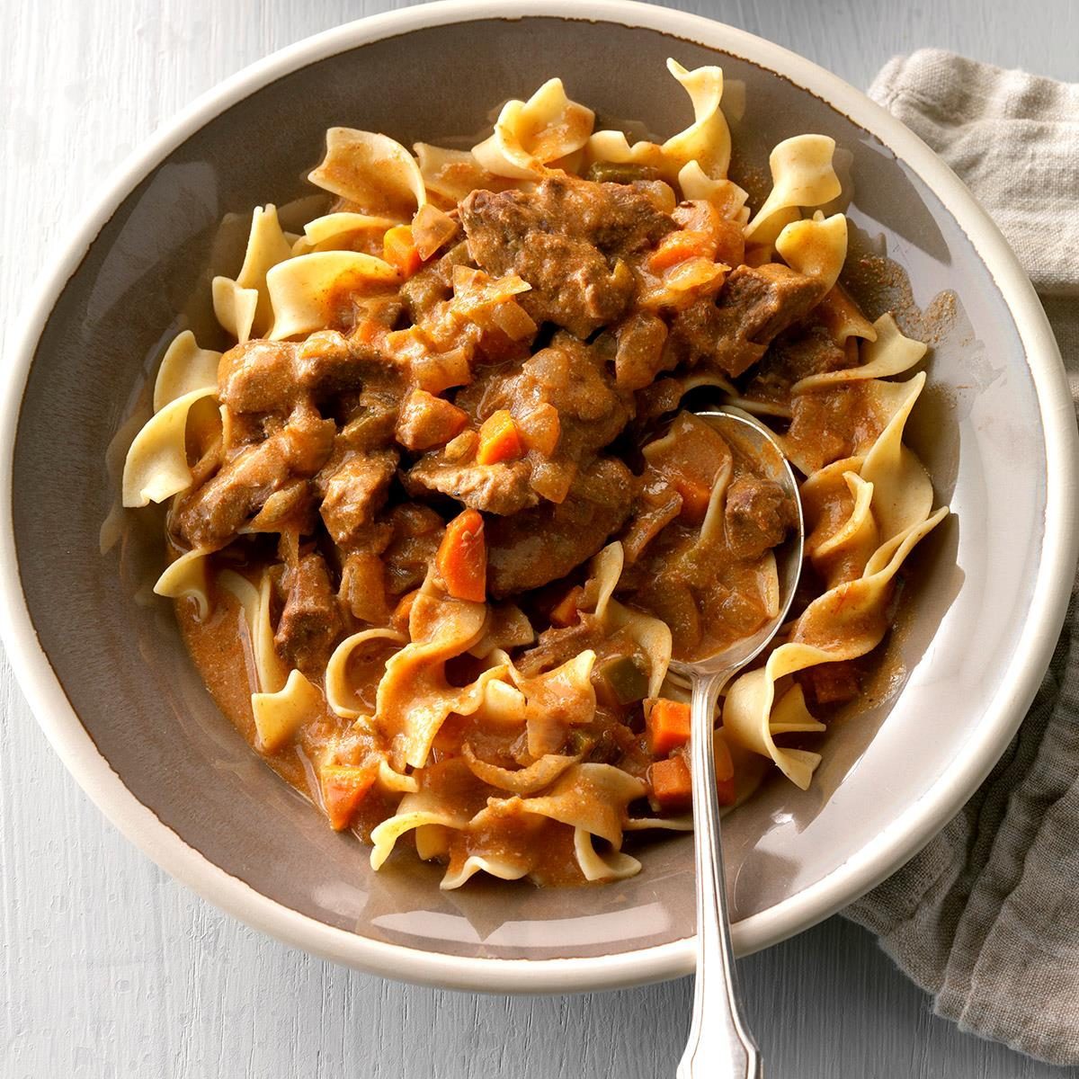 Pasta sits beneath beef stew with carrots in a bowl, accompanied by a silver spoon on a light surface with a cloth nearby.