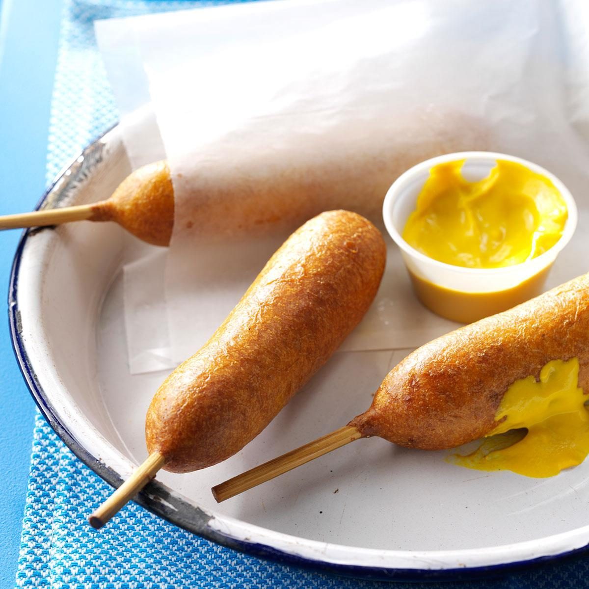 Corn dogs rest on a white plate, next to a small cup of yellow mustard, over a blue woven placemat.