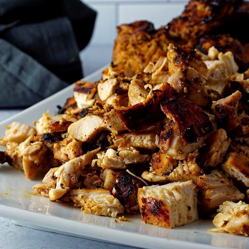 Grilled chicken pieces rest on a white plate, nestled in a kitchen setting with a dark cloth nearby.