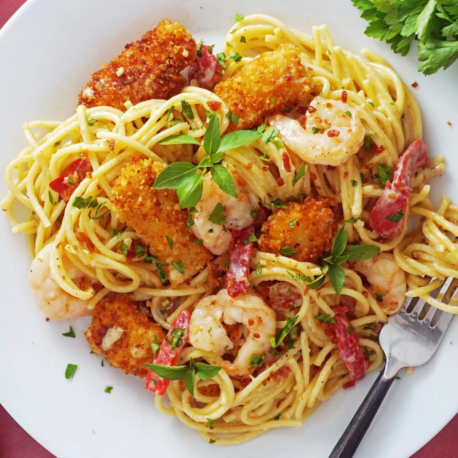 Pasta intertwined with shrimp and fried chicken pieces, garnished with herbs, on a white plate, accompanied by a fork and fresh parsley leaves nearby.