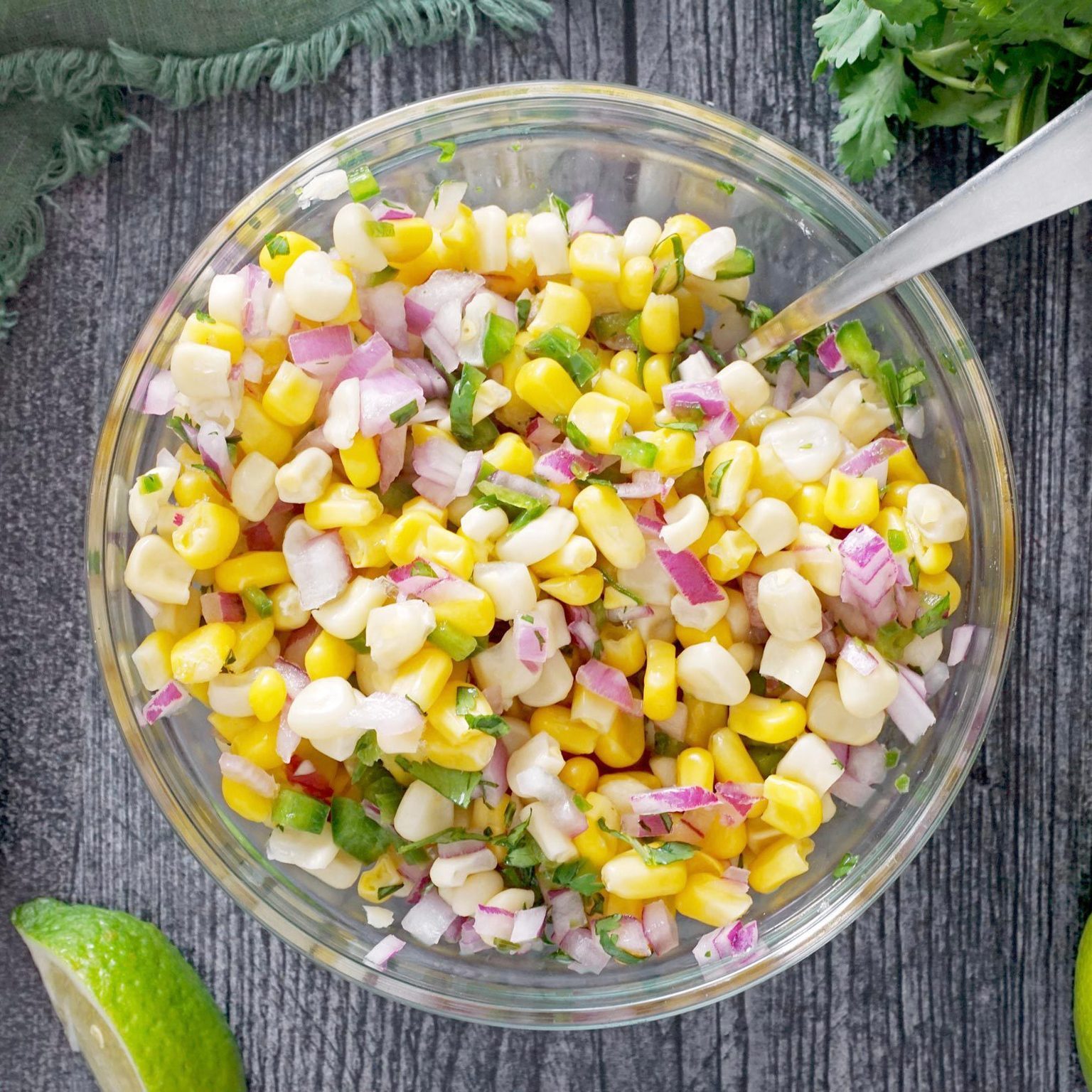 A glass bowl holds corn salad with red onion and cilantro, placed on a textured wooden table beside lime slices and green herbs.